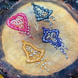 May include: Four embroidered Christmas ornaments in the shapes of a heart, a bell, and two Christmas trees. The ornaments are decorated with white beads and have a rustic, vintage look.