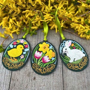 May include: Three green felt Easter egg ornaments with embroidered designs. The first egg features a yellow chick with the text "Frohe Ostern". The second egg features a bouquet of yellow daffodils with the text "Frohe Ostern". The third egg features a white rabbit with the text "Frohe Ostern".