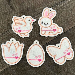 May include: Five felt Easter ornaments with pink and orange embroidery. The ornaments are shaped like a bird, bunny, tulip, egg, and butterfly.