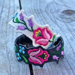 Op de afbeelding: Twee geborduurde viltarmbanden met bloemenmotieven. De ene armband is wit met roze, paarse en groene bloemen. De andere armband is zwart met roze, blauwe en groene bloemen.