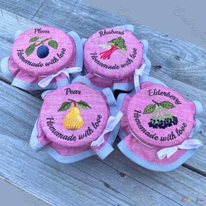 Op de afbeelding: Vier roze potdeksels met geborduurde fruitontwerpen: pruim, rabarber, peer en vlierbes. Elke deksel heeft de woorden "Homemade with love" geborduurd rond de fruitafbeelding. De deksels zijn afgewerkt met lichtblauwe stof en vastgebonden met witte linten.