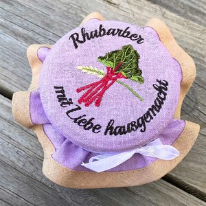 May include: A purple fabric jar lid with a brown trim and a white ribbon. The lid is embroidered with the text "Rhabarber mit Liebe hausgemacht" and a red and green rhubarb illustration.