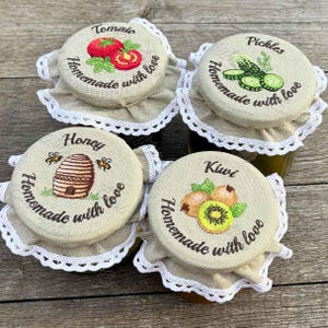 Puede incluir: Cuatro tarros de vidrio con tapas de tela bordadas. Las tapas presentan ilustraciones de tomates, pepinillos, miel y kiwi. Cada tapa está bordada con las palabras "Homemade with love" y ribeteada con encaje blanco. Los tarros están sobre una superficie de madera.