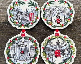 May include: Four embroidered Christmas ornaments with winter scenes. Each ornament features a different illustration of a snowy street scene, framed by a wreath of green pine needles and red berries. A red bow tops each ornament.