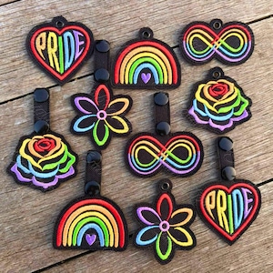 May include: A collection of embroidered patches with various designs, including rainbows, flowers, infinity symbols, and hearts. The patches feature vibrant rainbow colors and the word "PRIDE" on heart-shaped patches. Each patch has a black strap.
