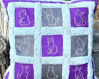 May include: A purple and gray pillow with a patchwork design featuring white embroidered cats in various poses. The pillow is made of a soft, fluffy fabric.