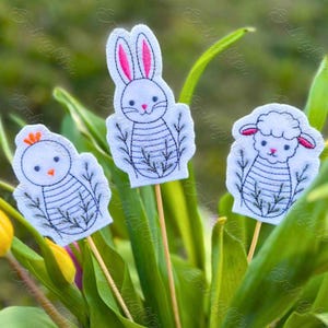 May include: Three felt Easter decorations on wooden sticks. A white bunny with pink ears, a white chick with an orange beak, and a white lamb with pink ears. Each animal has black and white stripes and is surrounded by green leaves.