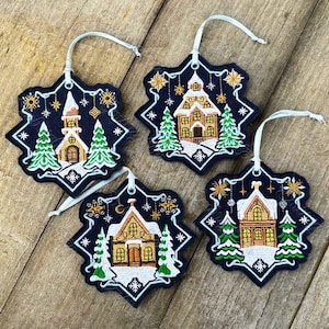 May include: Four snowflake-shaped Christmas ornaments with embroidered images of houses and trees. The ornaments are black with white and gold accents.
