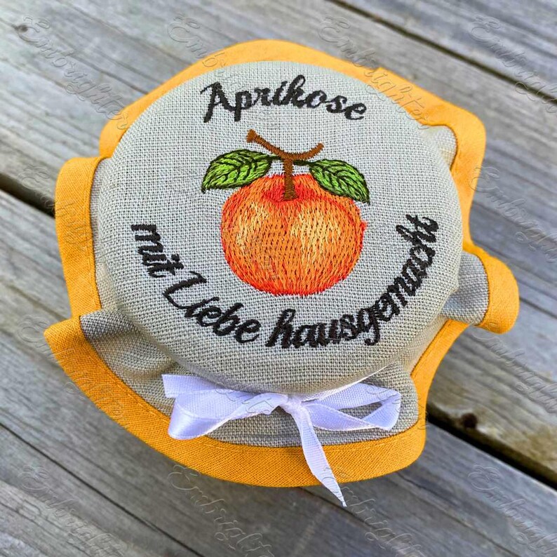 May include: Embroidered jar lid featuring an orange apricot with green leaves and the German text "Aprikose mit Liebe hausgemacht." The lid is gray with an orange border and a white ribbon bow.