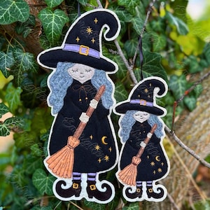 May include: Two felt witch ornaments featuring blue hair, black pointed hats with purple bands, and black dresses. Each witch holds a brown broomstick and is decorated with yellow embroidered stars and crescent moons, finished with black boots.