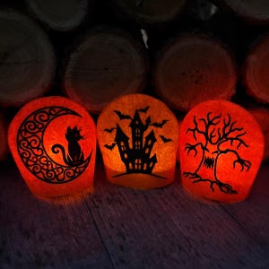 May include: Three orange, glowing, rounded decorative lights. The left light features a black cat sitting on a crescent moon. The center light displays a haunted house with flying bats. The right light shows a gnarled, spooky tree silhouette.