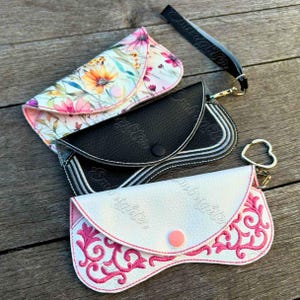 May include: Three eyeglass cases in a row. The top case has a floral pattern with pink, orange, and yellow flowers. The middle case is black with white stitching. The bottom case is white with pink embroidery and a pink button. A black wrist strap is attached to the top case.