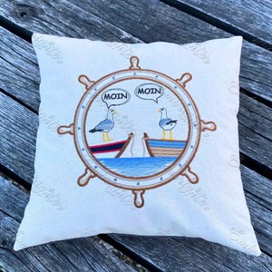 May include: A white pillow with a brown embroidered nautical wheel design. Two seagulls are perched on boats inside the wheel, with speech bubbles that say "Moin".