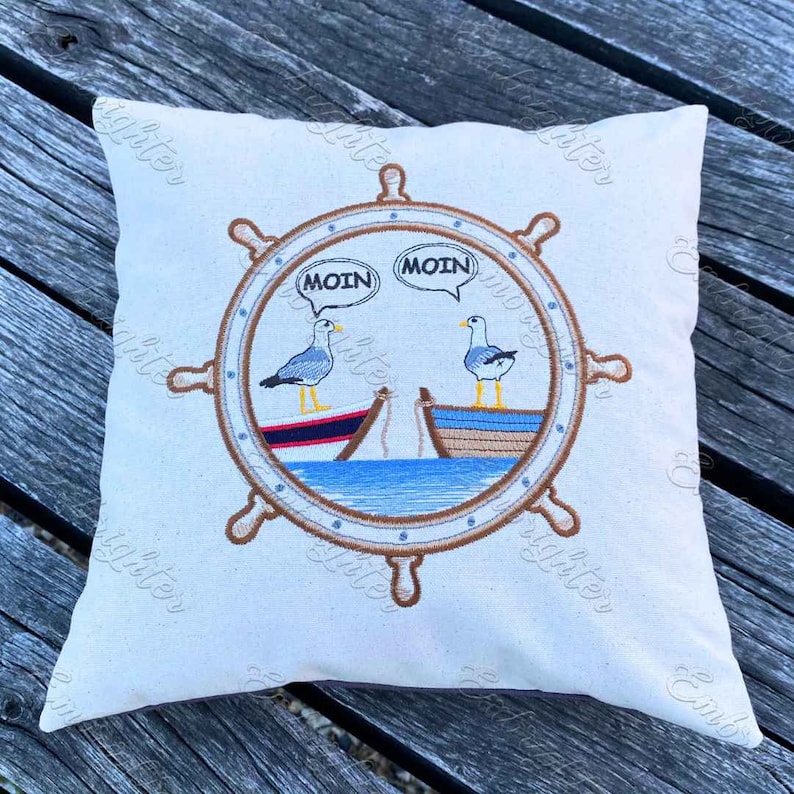 May include: A white pillow with a brown embroidered nautical wheel design. Two seagulls are perched on boats inside the wheel, with speech bubbles that say "Moin".