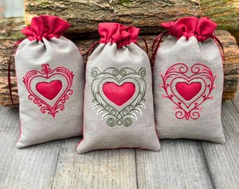 May include: Three small grey linen drawstring bags with red trim. Each bag has an embroidered red heart with a green and white floral design.
