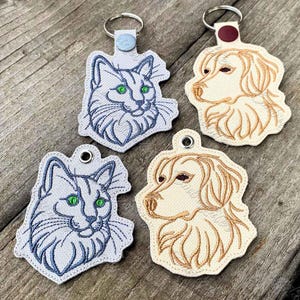 May include: Four embroidered keychains featuring animal portraits. Two keychains show a cat with blue stitching and green eyes, while two show a dog with tan stitching. Each keychain has a metal ring and a small snap button at the top.