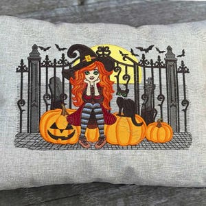 May include: Embroidered Halloween pillow featuring a witch with red hair, a black cat, and pumpkins. The design includes a black gate, bats, and a full moon. The color palette includes orange, black, and gray.