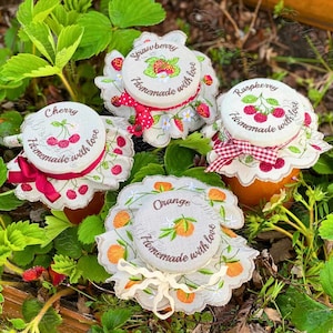 Può includere: Quattro vasetti di marmellata fatta in casa con coperchi ricamati. I coperchi sono decorati con diversi frutti: fragole, lamponi, ciliegie e arance. Ogni coperchio ha il nome del frutto e il testo "Fatto in casa con amore".