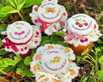 May include: Four jars of homemade jam with embroidered lids. The lids are decorated with different fruits: strawberries, raspberries, cherries, and oranges. Each lid has the fruit name and the text "Homemade with love".