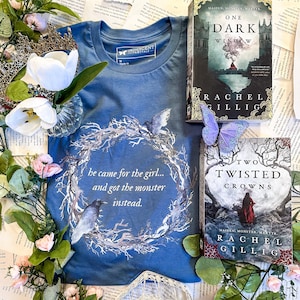 Pu&ograve; includere: Una maglietta blu con un design a corona bianca e il testo "he came for the girl... and got the monster instead." Sono anche raffigurati due libri: "One Dark Window" e "Two Twisted Crowns" di Rachel Gillig.