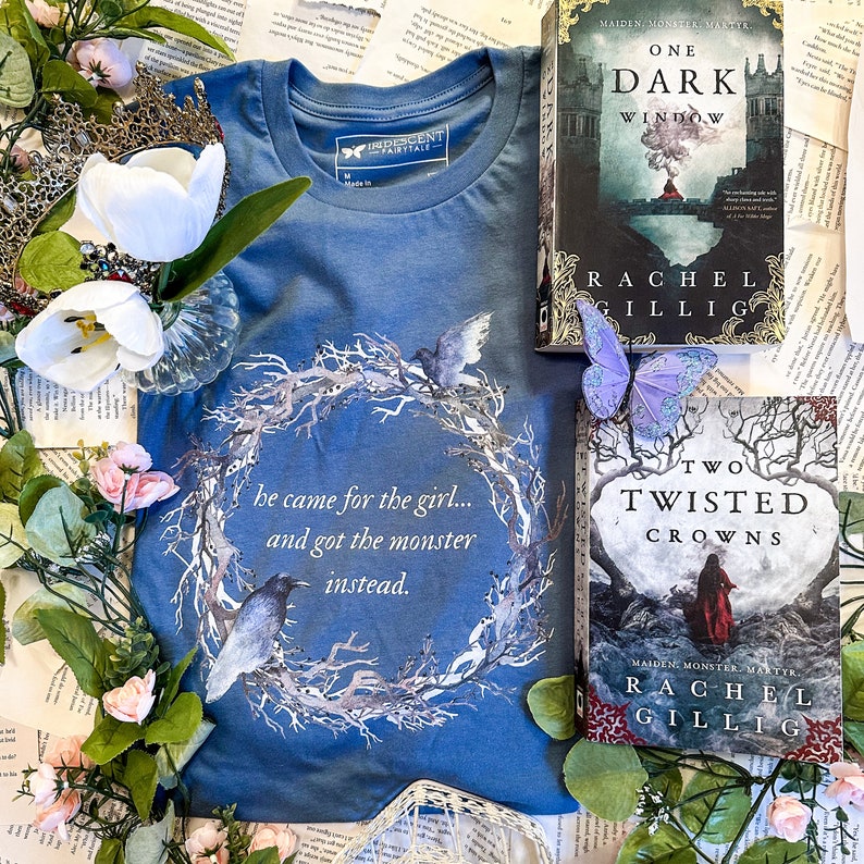Pu&ograve; includere: Una maglietta blu con un design a corona bianca e il testo "he came for the girl... and got the monster instead." Sono anche raffigurati due libri: "One Dark Window" e "Two Twisted Crowns" di Rachel Gillig.