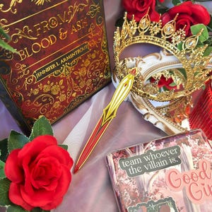 May include: A decorative scene featuring a book titled "FROM BLOOD & ASH" with gold lettering, a gold crown, a red and gold letter opener, and a sticker that says "team whoever the villain is."