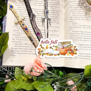 May include: A close-up of a book with a fall-themed sticker on it. The sticker features a stack of books, a pumpkin, and autumn leaves. The text on the sticker reads "hello fall".