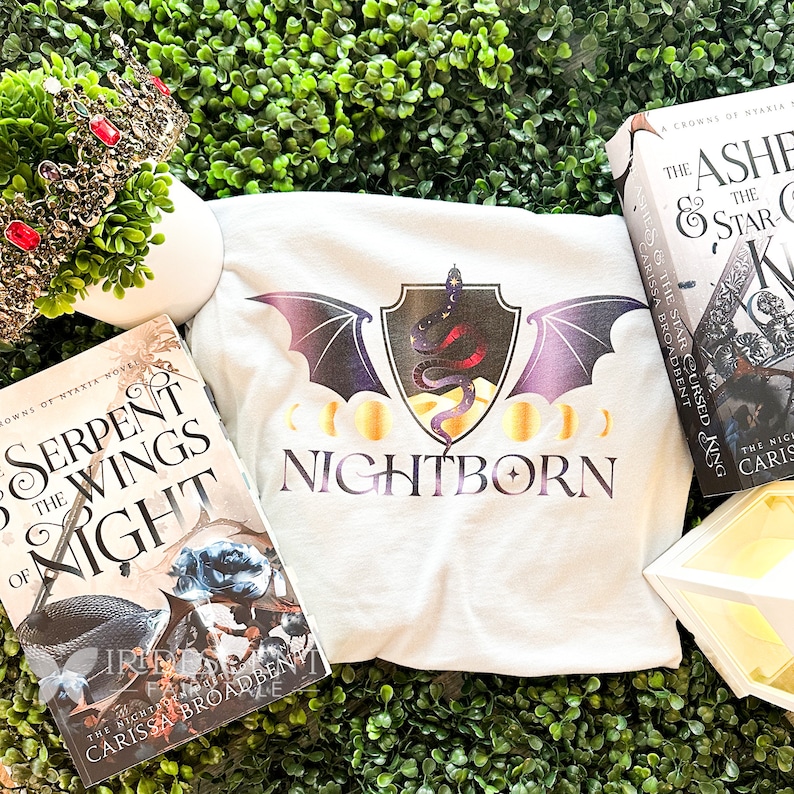 May include: A white t-shirt with a black and purple bat design and the text "NIGHTBORN" in a gold font. The bat has a gold shield with a snake on it. There are three gold moons on either side of the text. The t-shirt is lying on a green surface with a book titled "Serpent & The Wings of Night" and another book titled "The Ashes & The Star Cursed King" visible in the background.