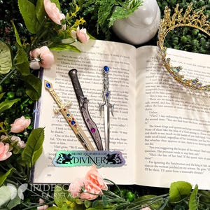 May include: Three decorative metal letter openers with jewel accents, resting on an open book with floral accents. A gold-colored crown is in the background. The text on the bookmark reads "THE ONLY GOD OF MEN-IS COIN DIVINER."