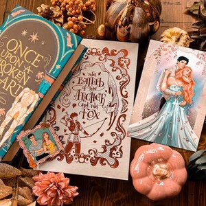 Puede incluir: Una disposici&oacute;n plana de tres libros con decoraci&oacute;n oto&ntilde;al. Los libros son "Once Upon a Broken Heart" de Stephanie Garber, "The Ballad of the Archer and the Fox" y un libro con una pareja en la portada. La decoraci&oacute;n incluye hojas de oto&ntilde;o, una peque&ntilde;a calabaza y una suculenta.