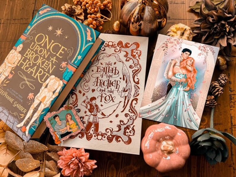 Puede incluir: Una disposici&oacute;n plana de tres libros con decoraci&oacute;n oto&ntilde;al. Los libros son "Once Upon a Broken Heart" de Stephanie Garber, "The Ballad of the Archer and the Fox" y un libro con una pareja en la portada. La decoraci&oacute;n incluye hojas de oto&ntilde;o, una peque&ntilde;a calabaza y una suculenta.