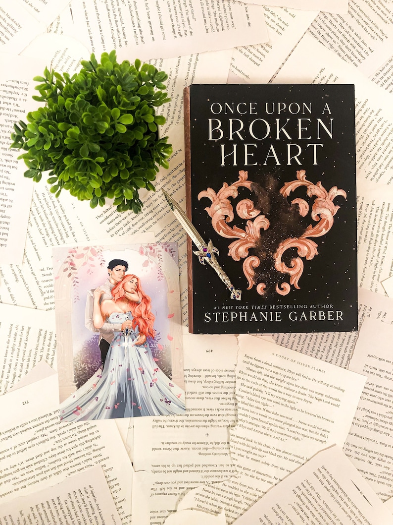 Puede incluir: Un libro titulado "Once Upon a Broken Heart" de Stephanie Garber, autora n&uacute;mero uno de bestsellers del New York Times. La portada del libro presenta un dise&ntilde;o en forma de coraz&oacute;n con un patr&oacute;n de galaxia y un marco dorado. El libro est&aacute; sobre una mesa con otros libros y una peque&ntilde;a planta verde.