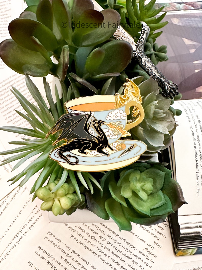 K&ouml;nnte beinhalten: Eine goldfarbene Emaille-Anstecknadel mit einer Teetasse mit Drachenmotiv. Die Teetasse ist blau und wei&szlig; mit einem goldenen Rand. Der Drache ist schwarz und hat eine goldene Umrandung. Die Teetasse ist mit einer hellorangen Fl&uuml;ssigkeit gef&uuml;llt. Die Anstecknadel ist vor einem Hintergrund aus gr&uuml;nen Sukkulenten platziert.