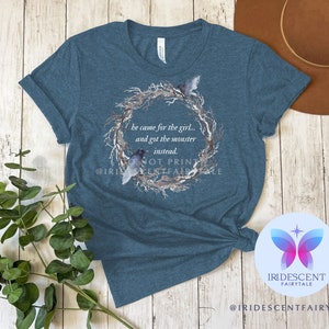 Pu&ograve; includere: Maglietta blu turchese con un design ad acquerello di una ghirlanda con uccelli e il testo "&Egrave; venuto per la ragazza... e si &egrave; preso il mostro invece." Il testo "Non stampare" e l'account social "@IRIDESCENTFAIRYTALE" sono visibili anche sulla maglietta.