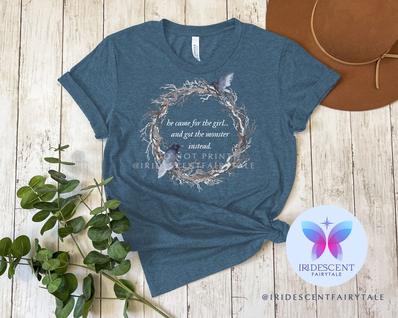 Pu&ograve; includere: Maglietta blu turchese con un design ad acquerello di una ghirlanda con uccelli e il testo "&Egrave; venuto per la ragazza... e si &egrave; preso il mostro invece." Il testo "Non stampare" e l'account social "@IRIDESCENTFAIRYTALE" sono visibili anche sulla maglietta.