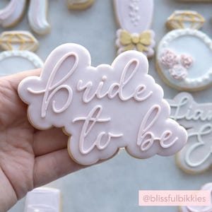 Op de afbeelding: Een lichtroze koekje in de vorm van een wolk met de woorden "Bride to be" in cursief geschreven. Andere koekjes op de achtergrond zijn een diamanten ring, een strik en een champagneglas. Het koekje wordt in een hand gehouden.