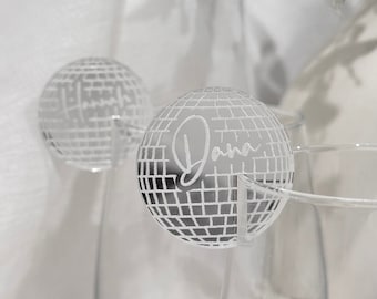 May include: Clear glass wine glasses with a decorative disco ball-shaped name tag that reads "Dana" in a cursive font. The disco ball is silver and has a grid pattern. The background is white.