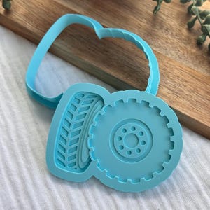 May include: A light blue plastic cookie cutter shaped like a tire with a detailed tread pattern and a central wheel rim design. The tool features a raised outer edge for cutting and an inner embossed section to imprint the tire texture into dough.