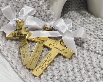 May include: Gold-colored cross favors with white ribbon bows. Each cross is engraved with the name "Frankie" and the text "Christening 27.08.2023". The crosses are displayed on a gray knitted surface.