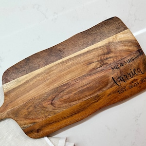 May include: A wooden serving board with a handle, featuring a natural wood grain pattern. The board is engraved with "MR & MRS Varriga EST. 26.03.2022" in a stylish font. The board rests on a white surface with a folded white cloth.