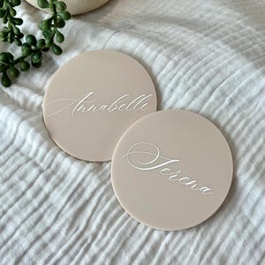 May include: Two round, beige place cards with white cursive text. One reads "Anabelle" and the other "Serena". The cards are on a white, textured surface, with a plant in the background.