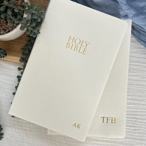 May include: Two off-white hardcover books with gold foil lettering. The top book features the text HOLY BIBLE centered and the initials AK in the bottom right corner. The bottom book features the initials TFB in the bottom right corner.