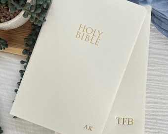 May include: Two off-white hardback books with gold foil lettering. The top book features the text HOLY BIBLE centred and the initials AK in the bottom right corner. The bottom book features the initials TFB in the bottom right corner.