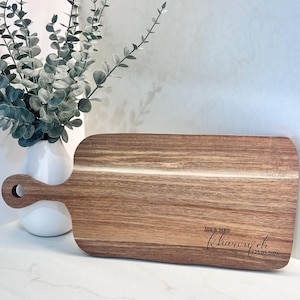 May include: A rectangular wooden cutting board with a handle and a hole for hanging. The board has a natural wood grain pattern and features the engraved text "MR & MRS Kharouyeh 23.05.2026".