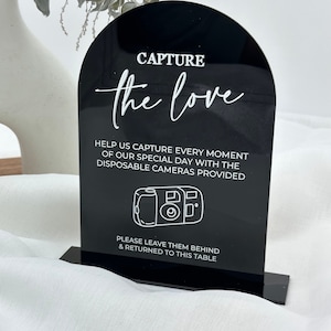 May include: Black acrylic wedding sign with an arched top. White text reads "CAPTURE the love" and instructions for disposable cameras. A line drawing of a camera is below the text. The sign is on a black base.