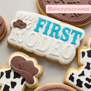 May include: Decorated sugar cookies with a rodeo theme. One cookie has the text "FIRST RODEO" in blue and white icing, with a cowboy hat. Other cookies feature cowboy hats and cowhide patterns. The cookies are on a light background.