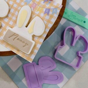 Peut inclure: Des biscuits de Pâques en forme de têtes de lapin avec des noms, disposés sur une assiette avec des œufs de Pâques et un fond à carreaux vichy. Les biscuits ont des accents roses et jaunes.