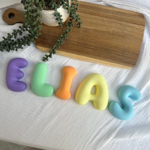 colorful letters spelling out the name "Elias" on a white surface, with a wooden cutting board and a potted plant in the background.