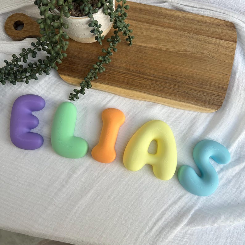 colorful letters spelling out the name "Elias" on a white surface, with a wooden cutting board and a potted plant in the background.