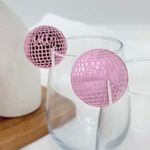 May include: Two pink disco ball-shaped drink charms with names, "Polly" and "Kara", etched on them. The charms are designed to sit on the rim of a glass. The background is a blurred white and light gray.